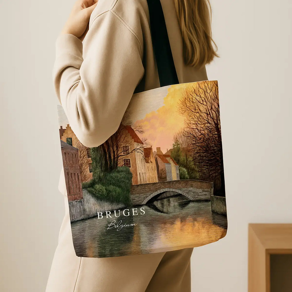 Person holding a tote bag with a scenic design of Bruges, featuring a bridge over water and buildings.