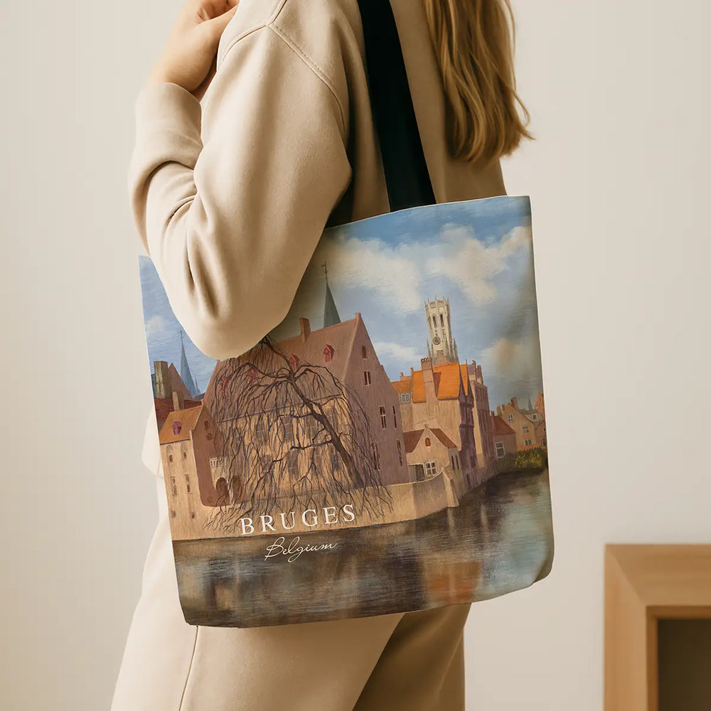 Person holding a tote bag with a scenic design of Bruges, featuring buildings and water.