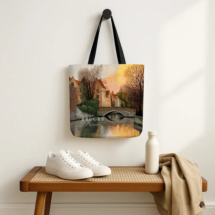 Tote bag with a scenic design of Bruges hanging on a wall above a wooden bench with shoes and a bottle.