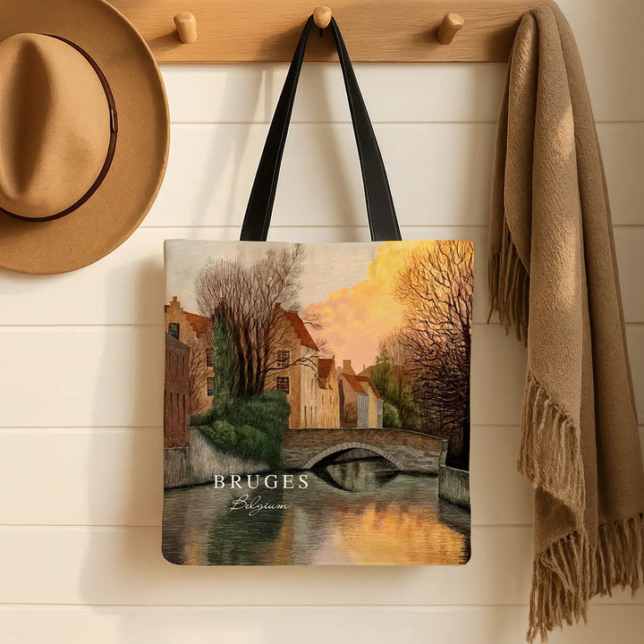Tote bag with a scenic design of Bruges, hanging on a wooden hook with a hat and scarf in the background.