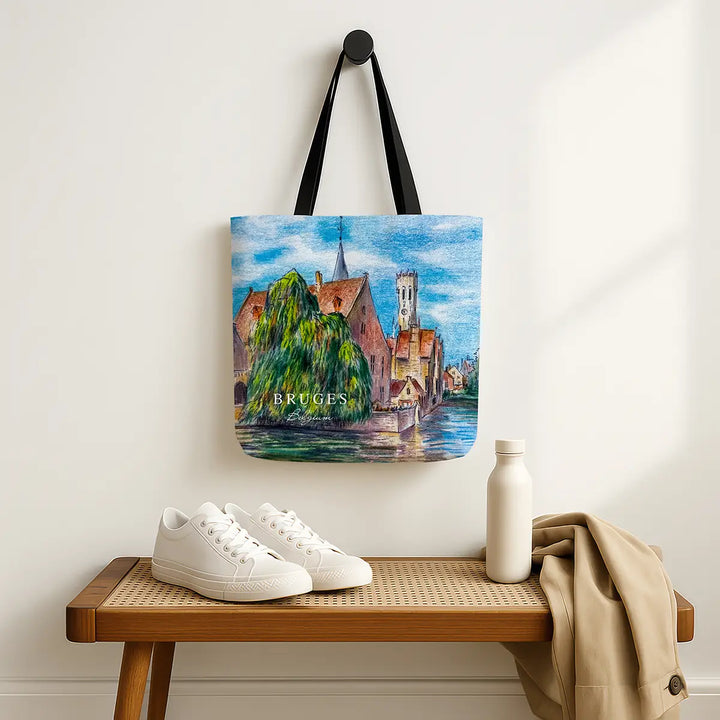 Tote bag with a scenic design of Bruges, Belgium, hanging on a wall above a wooden bench with shoes and a bottle.